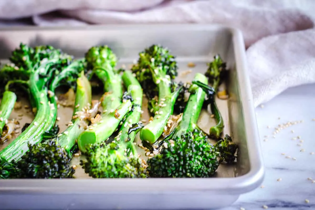 Closeup image of Roasted Broccoli with Sesame & Garlic. A silver baking tray of roasted broccoli is visible on a white marble background. Surrounding it are some sesame seeds and a linen napkin.