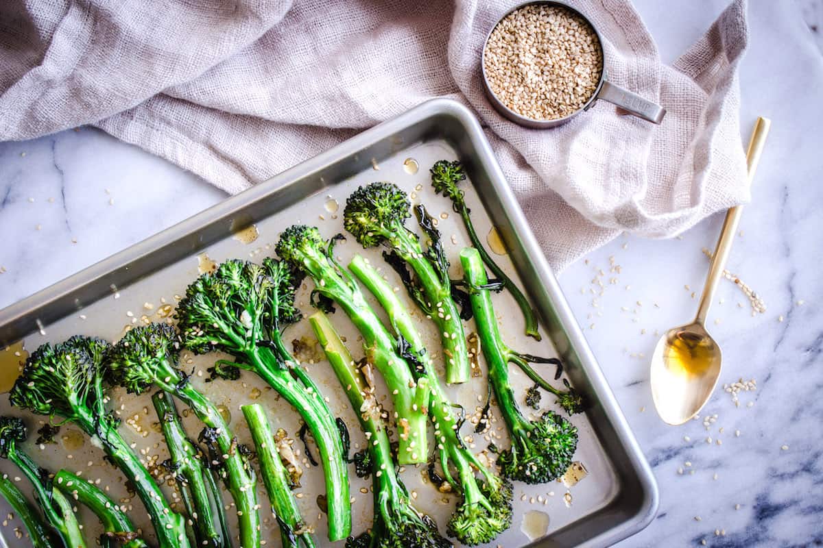 Overhead image of Roasted Broccoli with Sesame & Garlic. A silver baking tray of roasted broccoli is visible on a white marble background. Surrounding it is a golden spoon, a cup of sesame seeds and a linen napkin.