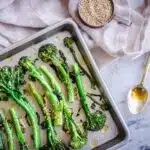 Overhead image of Roasted Broccoli with Sesame & Garlic. A silver baking tray of roasted broccoli is visible on a white marble background. Surrounding it is a golden spoon, a cup of sesame seeds and a linen napkin.