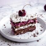 Slice of No-Bake Black Forest Cake with layers of cherry jam and coconut cream. The cake is decorated with a whole cherry and chocolate shavings. It is placed on a white plate on a white marble countertop, and is surrounded by silver cutlery, a cake slice, cherries and more chocolate shavings.