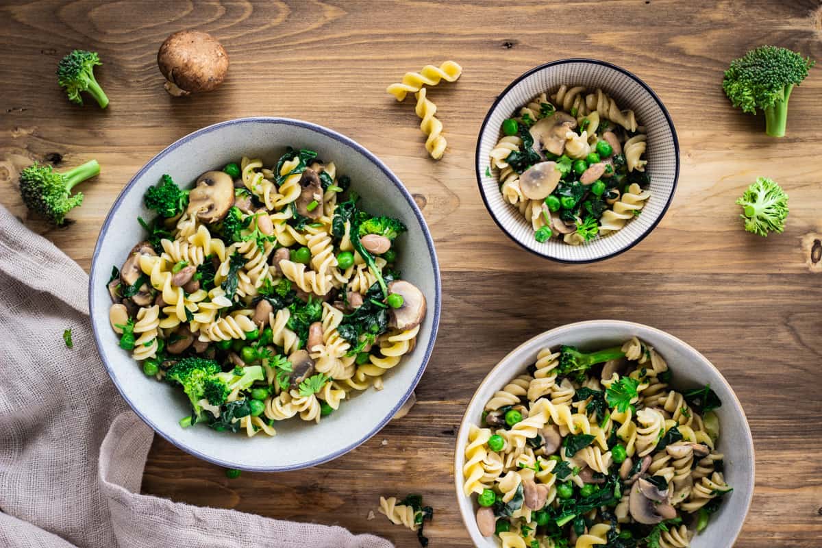 Image shows bowl of Creamy Pasta with Broccoli & Mushrooms on a wooden table. The bowl is surrounded by a linen napkin, bits of pasta and other bowls.