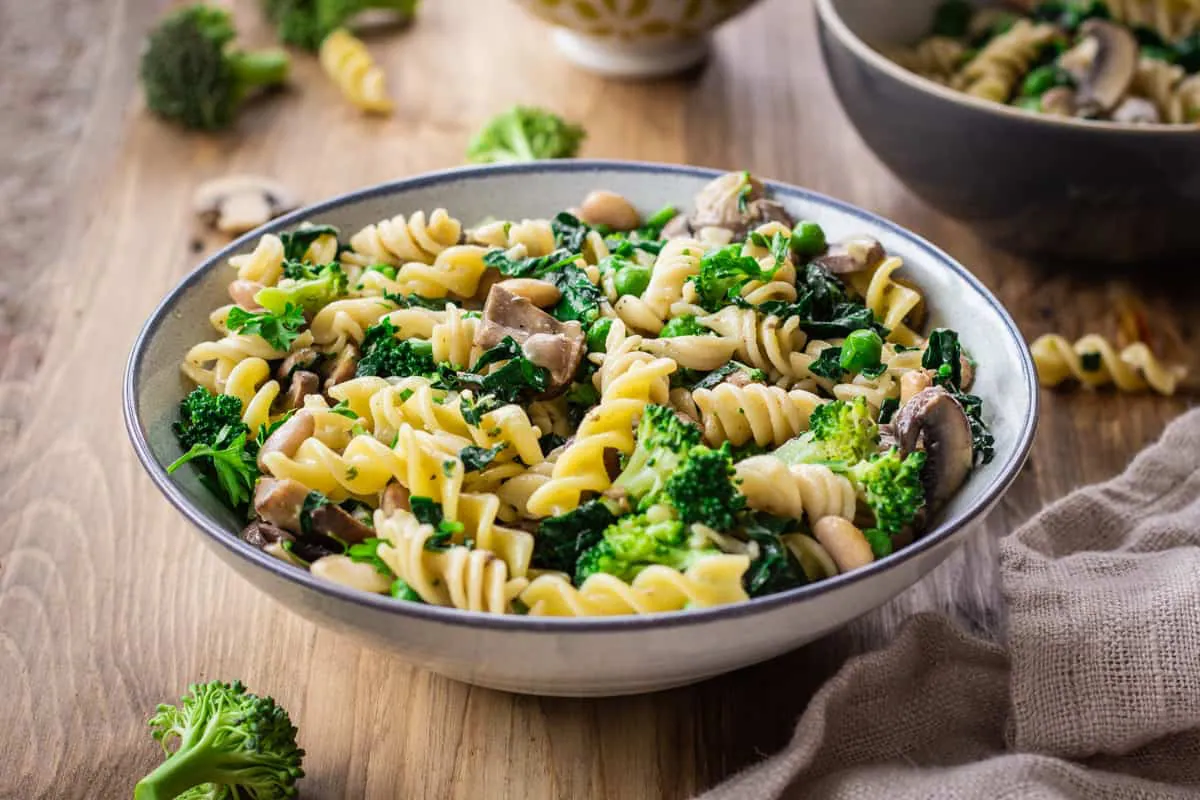 Image shows bowl of Creamy Pasta with Broccoli & Mushrooms on a wooden table. The bowl is surrounded by a linen napkin, bits of pasta and other bowls.