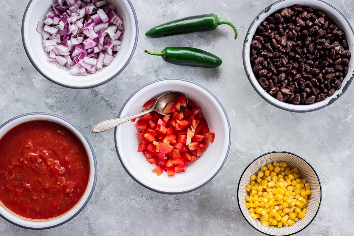 Image shows ingredients for Hearty Nacho Bake including bowls of chopped red onion, red pepper, sweet corn, salsa, black beans and jalepenos. The bake is made with Que Pasa Salted Tortilla Chips and Mexicana Mild Salsa.