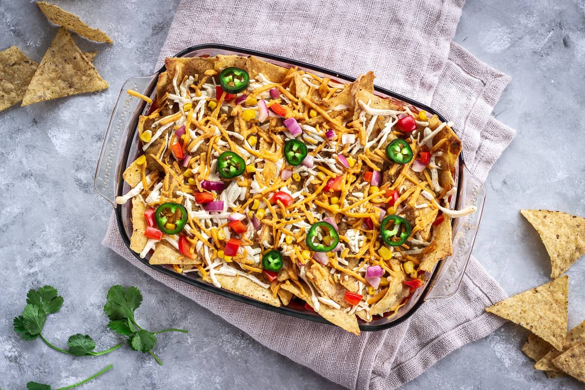 Overhead image shows Hearty Nacho Bake decorated with vegan cheese shreds and jalapenos. The bake is surrounded by scattered Que Pasa Salted Tortilla Chips and is made with their Mexicana Mild Salsa.