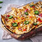 Image shows Hearty Nacho Bake decorated with vegan cheese shreds and jalapenos. The bake is surrounded by cilantro and scattered Que Pasa Salted Tortilla Chips, and is made with their Mexicana Mild Salsa.