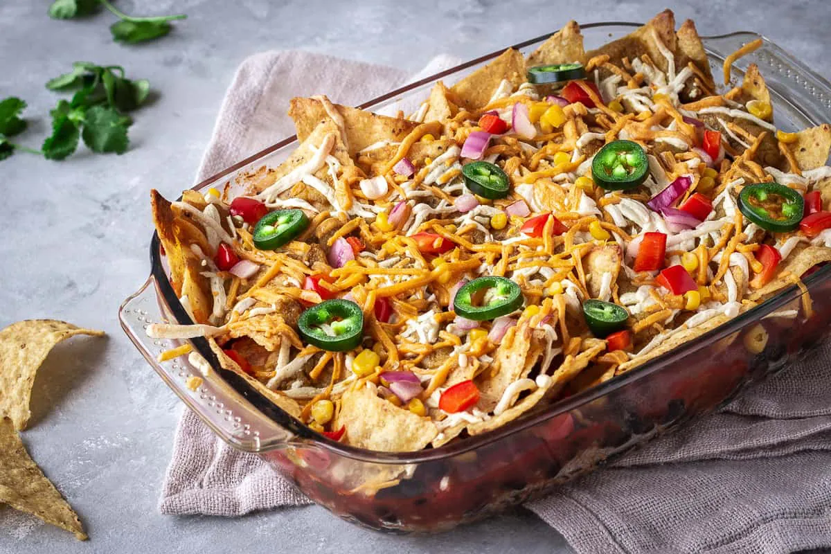 Image shows Hearty Nacho Bake decorated with vegan cheese shreds and jalapenos. The bake is surrounded by cilantro and scattered Que Pasa Salted Tortilla Chips, and is made with their Mexicana Mild Salsa.