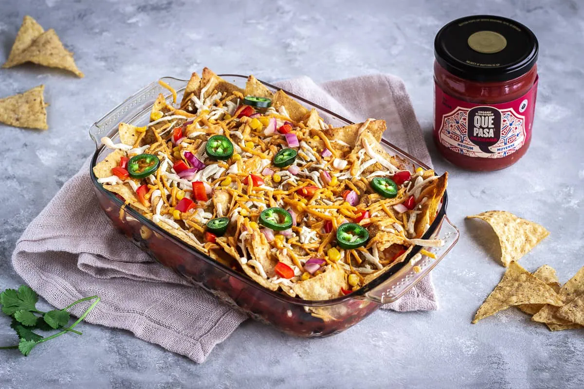 Image shows Hearty Nacho Bake decorated with vegan cheese shreds and jalapenos. The bake is surrounded by scattered Que Pasa Salted Tortilla Chips and a jar of their Mexicana Mild Salsa.