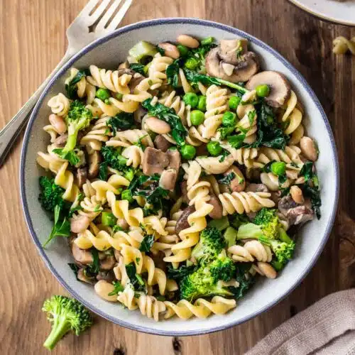 Image shows bowl of Creamy Pasta with Broccoli & Mushrooms on a wooden table. The bowl is surrounded by a silver cutlery, a linen napkin, bits of pasta and other bowls.