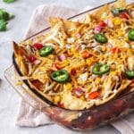 Hearty Nacho Bake decorated with vegan cheese shreds and jalapenos. The bake is surrounded by cilantro and scattered Tortilla Chips.