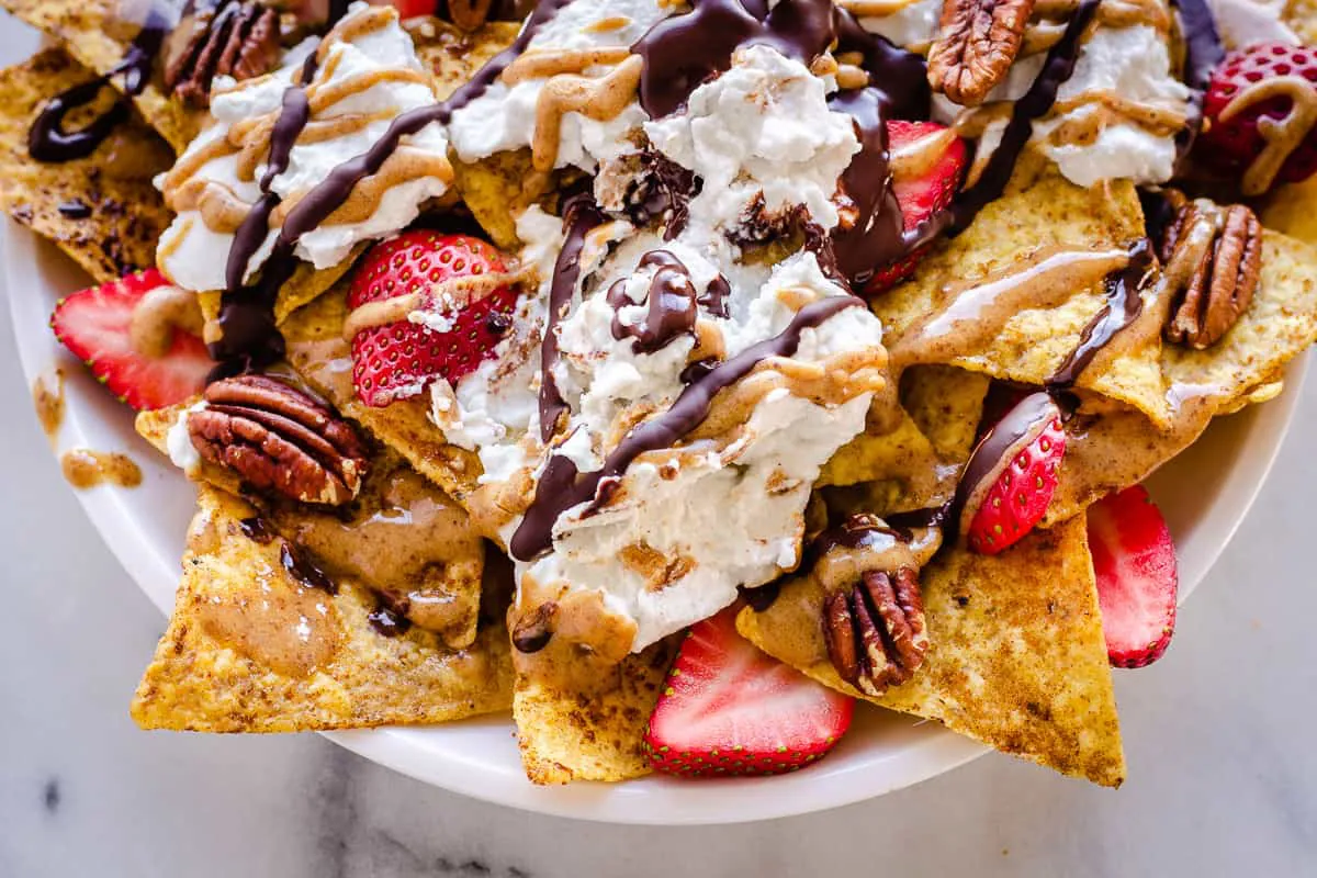 Close-up overhead image of Dessert Nachos . The nachos are decorated with strawberries, coconut whipped cream, pecans, drizzled chocolate and almond caramel sauce.
