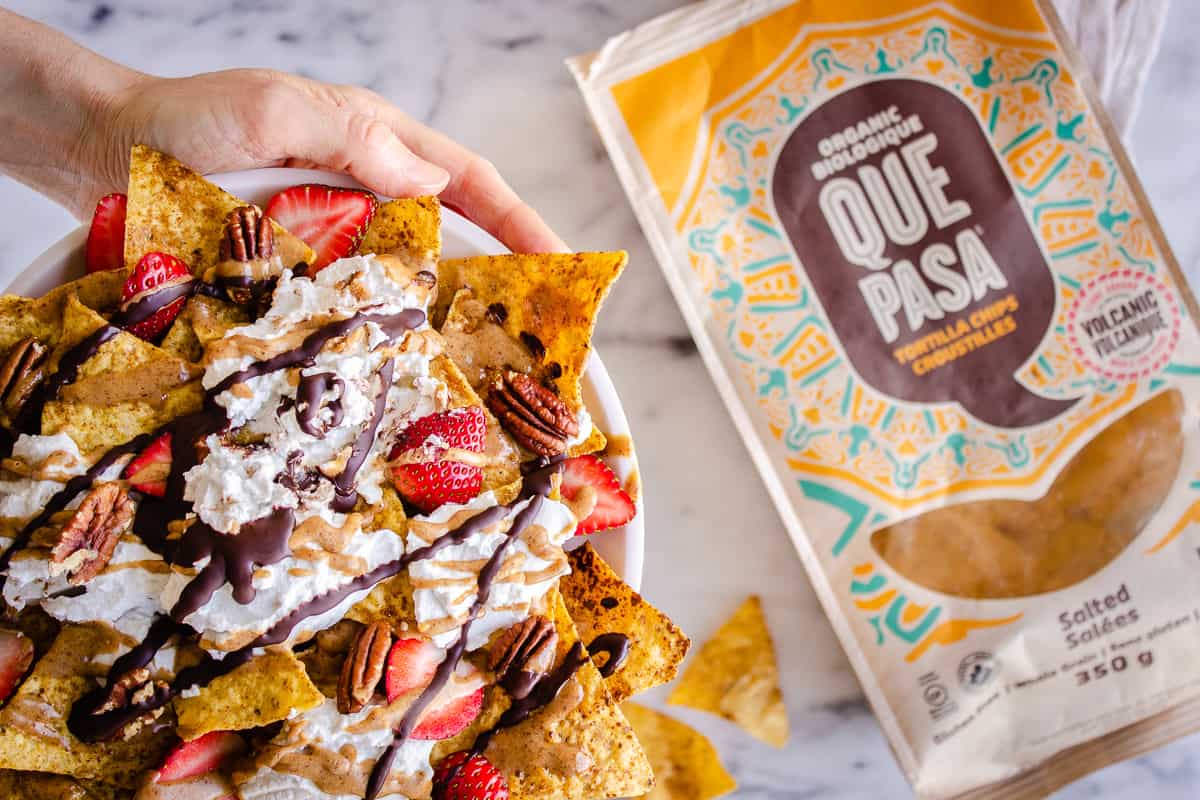 Overhead image of Dessert Nachos showing hands holding a plate of them and a Que Pasa tortilla chip bag in the background. The nachos are decorated with strawberries, coconut whipped cream, pecans and almond caramel sauce.