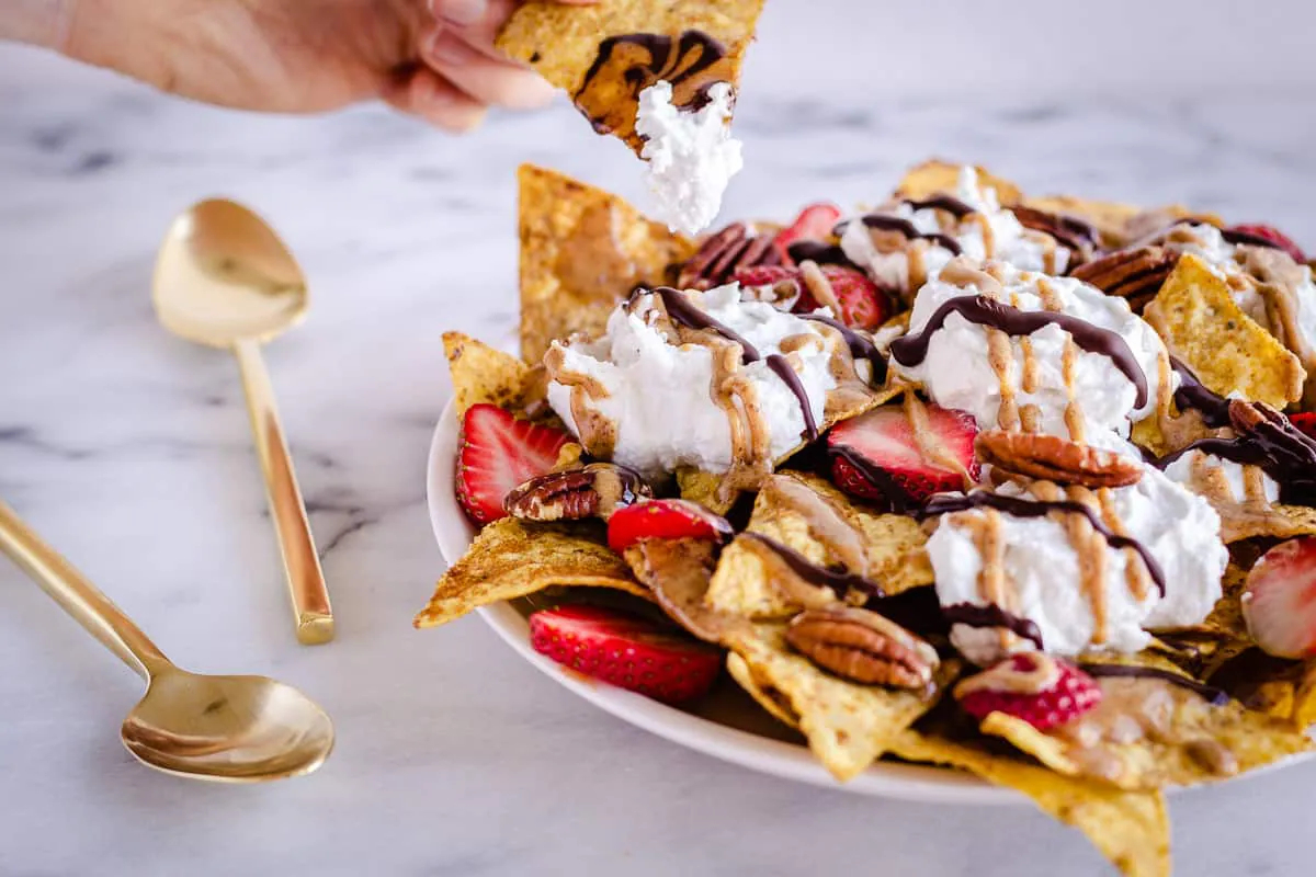 Image of Dessert Nachos showing a hand taking a nacho with cream and chocolate on. The nachos are decorated with strawberries, coconut whipped cream, pecans, drizzled chocolate and almond caramel sauce.