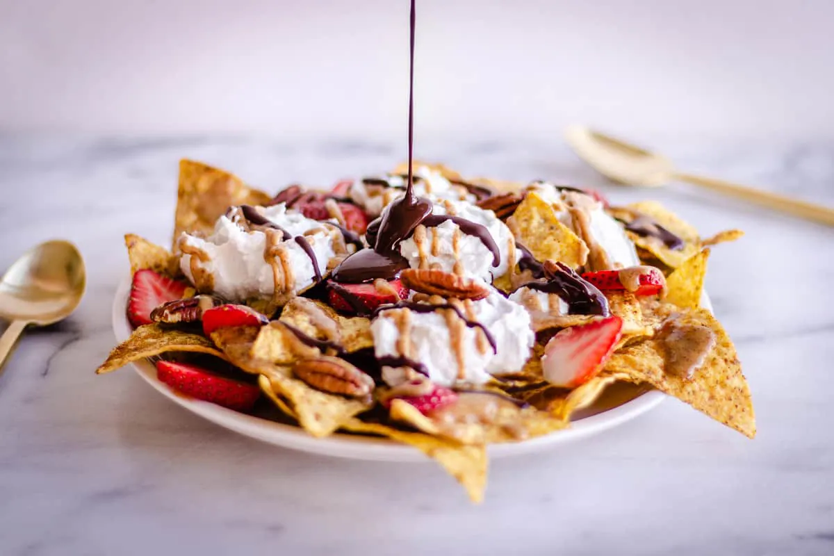 Image of Dessert Nachos showing dark chocolate being drizzled over them. The nachos are decorated with strawberries, coconut whipped cream, pecans, and almond caramel sauce.