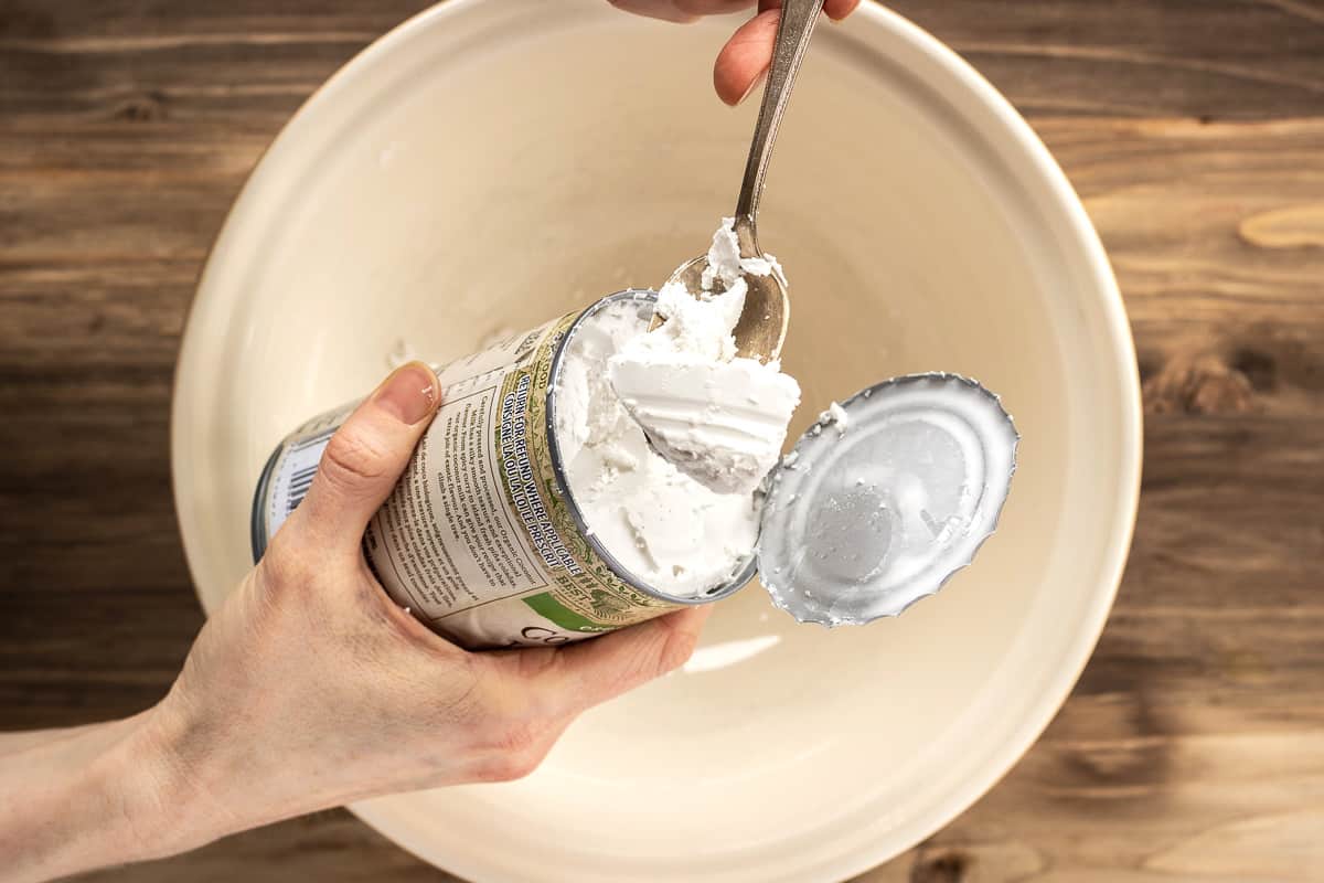 Image shows a woman's hands scraping hardened coconut cream from a can into a large mixing bowl with a silver spoon.