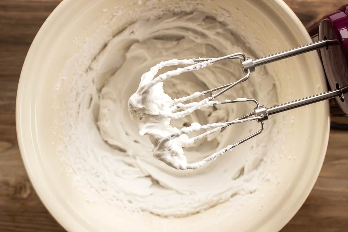 Image shows coconut whipped cream forming soft peaks on electric beaters. Behind the beaters is a bowl full of the cream.