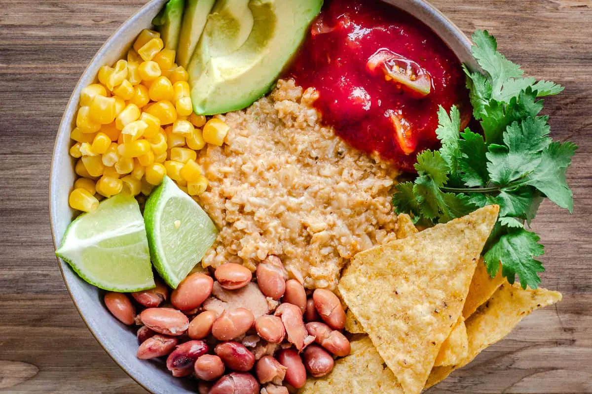 Close up overhead image of Savoury Vegan Oatmeal in large bowl on wooden background. Oatmeal is topped with avocado, corn, salsa, tortilla chips, lime, cilantro and beans.