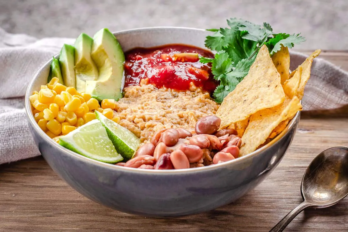 Close up image of Savoury Vegan Oatmeal in large bowl on wooden table. Oatmeal is topped with avocado, corn, salsa, tortilla chips, lime, cilantro and beans and a linen napkin is behind it.