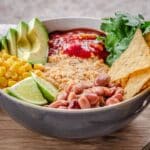 Savoury Vegan Oatmeal in large bowl on wooden table. Oatmeal is topped with avocado, corn, salsa, tortilla chips, lime, cilantro and beans and a linen napkin is behind it.