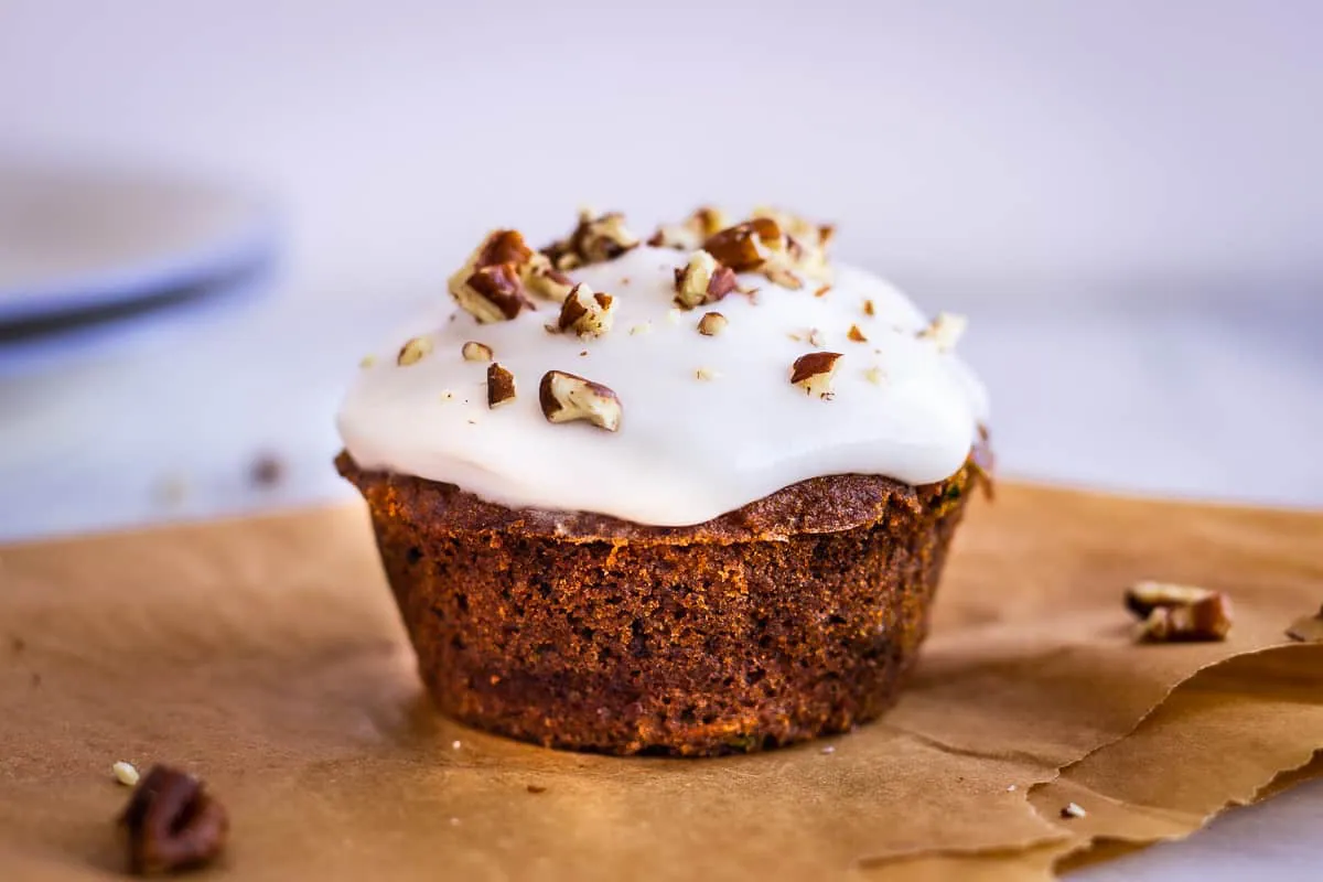 Close up image of Carrot Cake Muffins. Image shows a muffin on baking parchment, decorated with white frosting and pecans.