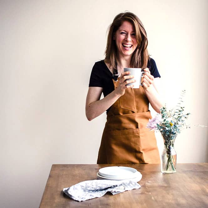 Image of Elizabeth Emery, food blogger at Vancouver with Love. Elizabeth is standing behind a wooden table wearing a brown apron and holding a mug of tea. There is a vase of wildflowers on the table, some plates and a grey napkin.