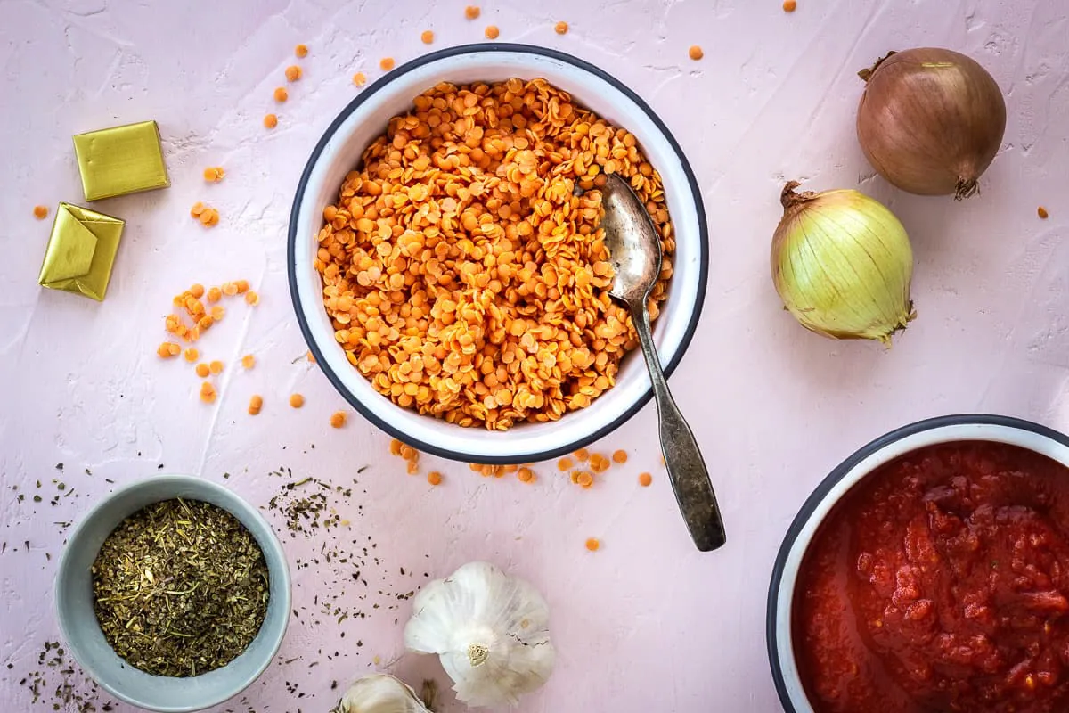 Overhead image of ingredients for Store Cupboard Lentil Bolognese on a light pink background. Image shows onions, garlic, red lentils, crushed tomatoes, dried herbs and stock cubes.