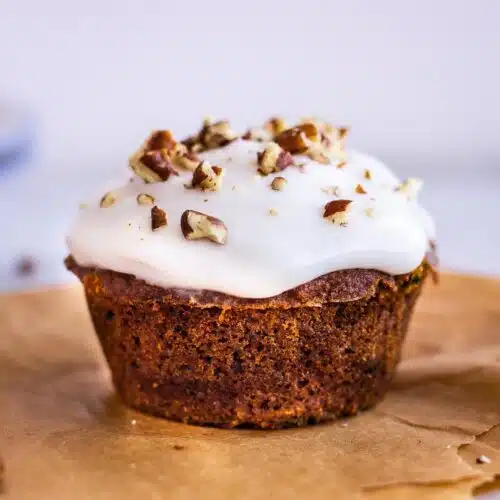 Close up image of Carrot Cake Muffins. Image shows a muffin on baking parchment, decorated with white frosting and pecans.
