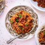 Image of several plates of lentil bolognese with spaghetti on a light pink surface. Two of the plates have forks stuck in the spaghetti.