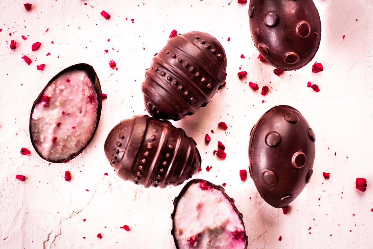 Image shows overhead view of patterned vegan chocolate eggs on a pale pink background. Some of the egg halves are filled with raspberry cream and dried raspberry pieces are scattered around the eggs.