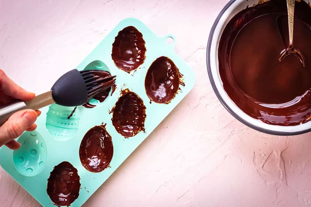 Image shows a woman's hand brushing melted chocolate into turquoise silicon egg moulds with a pastry brush. There is a bowl of melted chocolate nearby with a spoon.