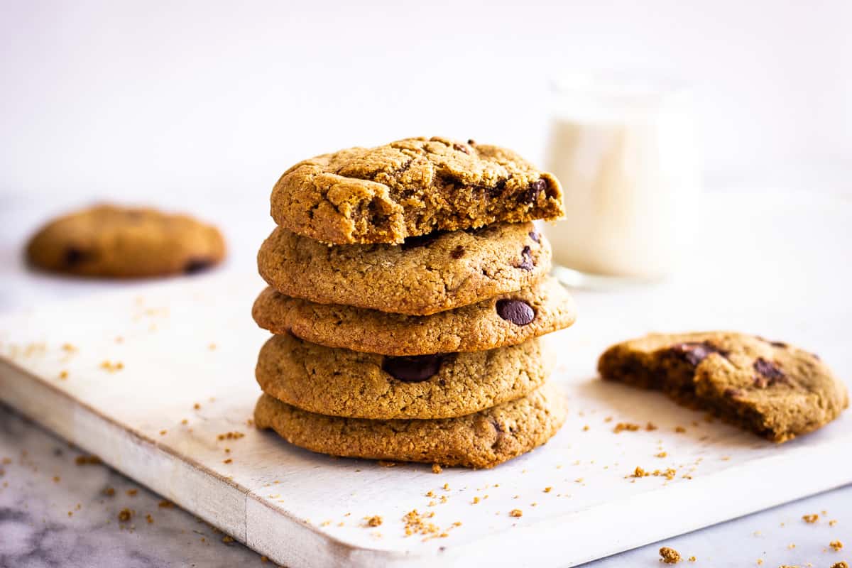 Close up image of Oatmeal Chocolate Chip Cookies. Cookies are on a white wooden chopping board on a marble background. Around them are some cookie crumbs and in the background is a glass of plant milk.
