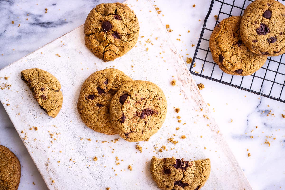 Overhead shot of Oatmeal Chocolate Chip Cookies. Cookies are on a white wooden chopping board on a marble background. Around them are some cookie crumbs and in the corner of the image is a wire cooling rack containing some more cookies.
