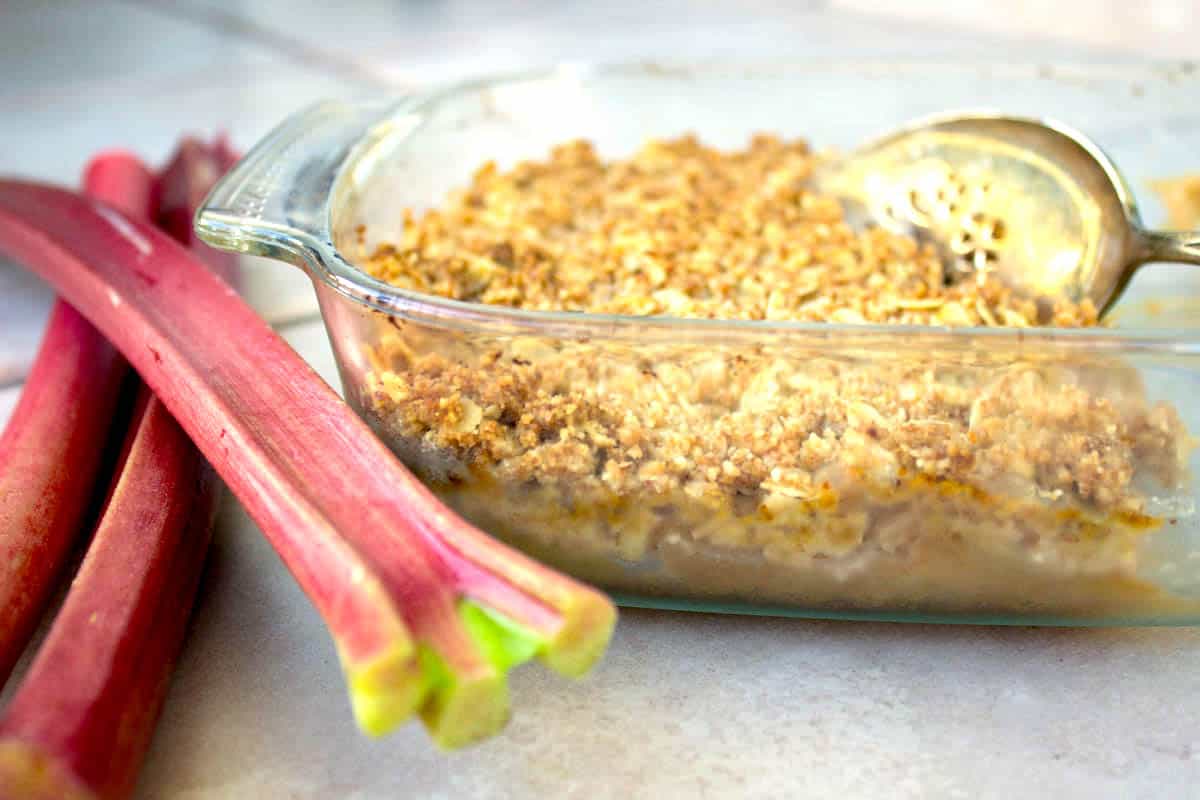 Close-up image of vegan Rhubarb & Apple Crumble. image shows crumble in a glass dish with a silver cake slice, surround by some stalks of rhubarb.