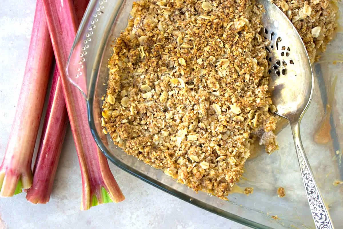 Overhead image of vegan Rhubarb & Apple Crumble. image shows crumble in a glass dish with a silver cake slice, surround by some stalks of rhubarb.