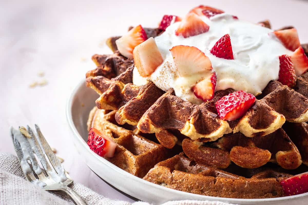 Closeup image of 3 Ingredient Oatmeal Waffles. A stack of waffles is on a grey plate on a light pink background. The waffles are decorated with strawberries and coconut whipped cream, and next to them is a silver spoon with cream on it and a linen napkin.