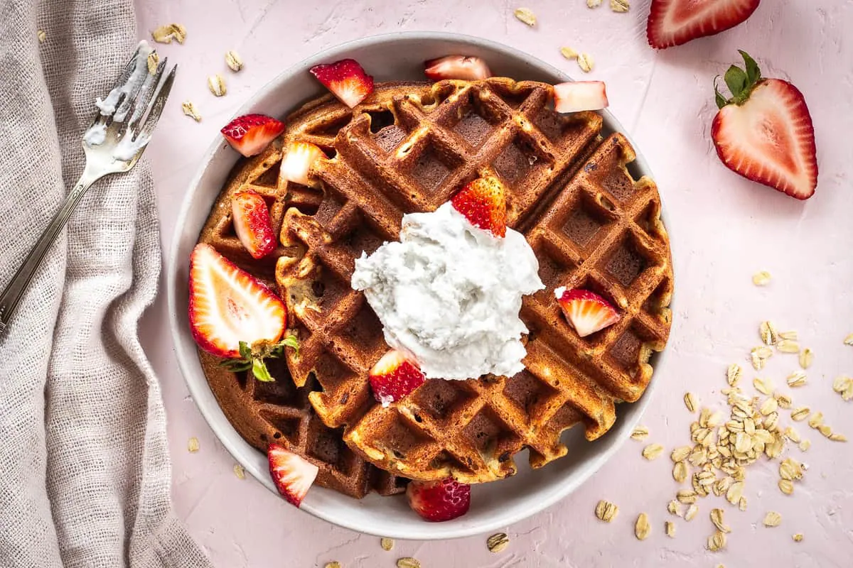 Overhead image of 3 Ingredient Oatmeal Waffles. A stack of waffles is on a grey plate on a light pink background. The waffles are decorated with strawberries and coconut whipped cream, and are surrounded by a silver spoon with cream on it, scattered oats, strawberry halves and a linen napkin.