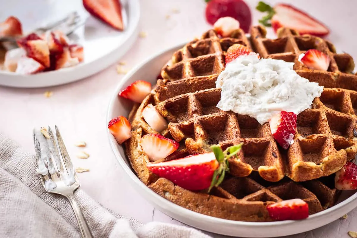 Close up image of 3 Ingredient Oatmeal Waffles. A stack of waffles is on a grey plate on a light pink background. The waffles are decorated with strawberries and coconut whipped cream, and are surrounded by a silver spoon with cream on it, scattered oats and a linen napkin. Another plate of strawberries is nearby.