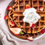 Overhead image of 3 Ingredient Oatmeal Waffles. A stack of waffles is on a grey plate on a light pink background. The waffles are decorated with strawberries and coconut whipped cream, and are surrounded by a silver spoon with cream on it, scattered oats and a linen napkin.