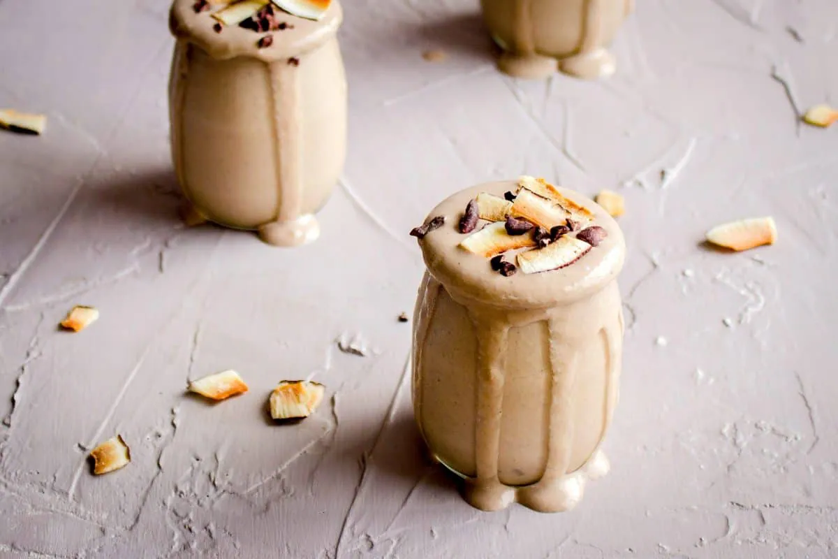Image of 5 Minute Post Workout Smoothie. Image shows close-up of cream-coloured smoothie in small glass, decorated with cacao nibs and coconut flakes. Two more smoothie glasses are visible in the background.