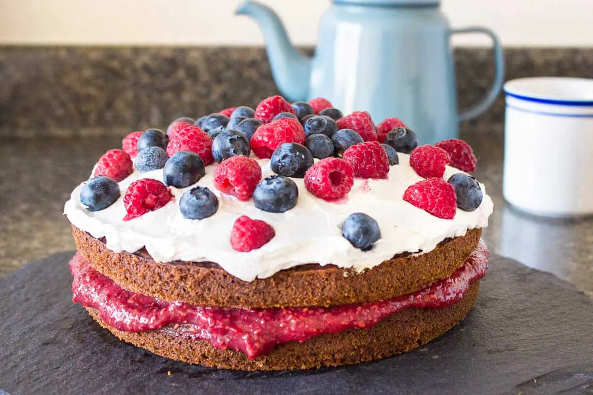 Vegan Gluten Free Sponge Cake. Cake is on a dark grey slate platter on a kitchen countertop and is decorated with raspberry jam, coconut whipped cream, blueberries and raspberries. In the background, a grey teapot and white striped cup are visible.