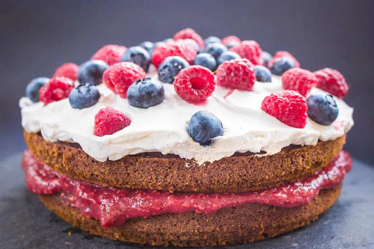 Vegan Gluten Free Sponge Cake. Cake is on a dark grey slate background and is decorated with raspberry jam, coconut whipped cream, blueberries and raspberries.