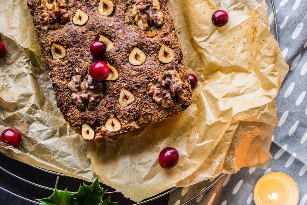 Overhead view of Classic Vegan Nut Roast. Nut roast is on parchment paper on a wire cooling rack, surrounded by a grey napkin, candles and holly. Some cranberries have been sprinkled over it.