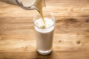 Image shows freshly made hemp milk being poured into a tall glass on a wooden table.