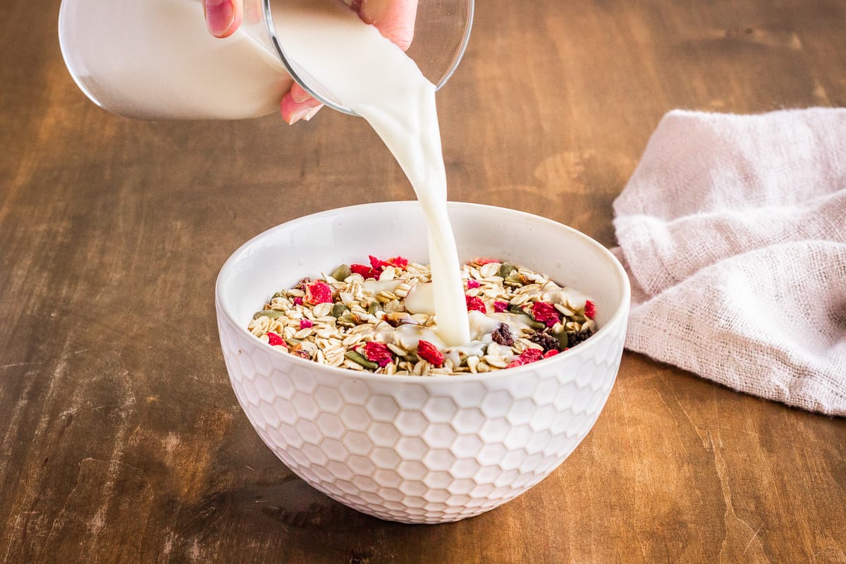 How to Make Hemp Milk - image shows a carafe of freshly made hemp milk being poured into a bowl of muesli. The bowl is on a wooden table and is surrounded by a linen napkin.