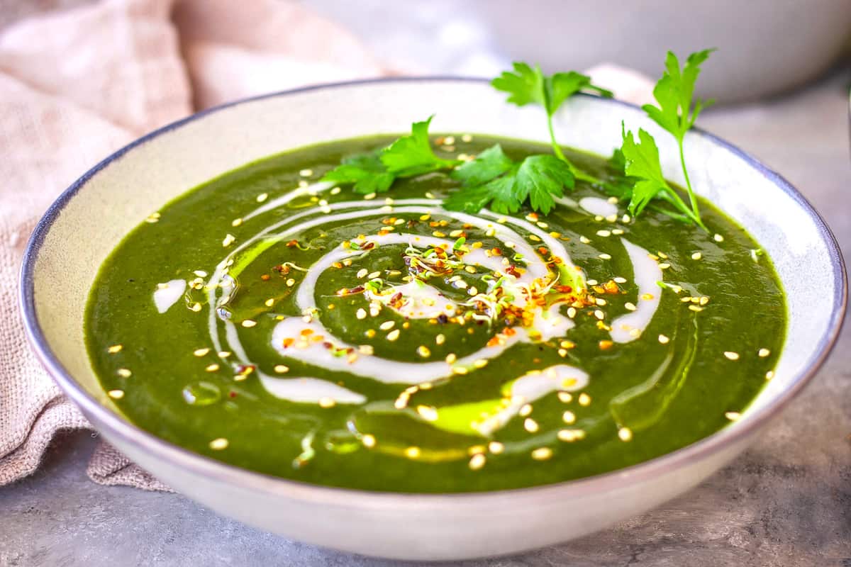 Close up image of a bowl of Nutrient-Rich Nettle Soup. The grey bowl is decorated with parsley leaves, yoghurt swirls and alfalfa sprouts.