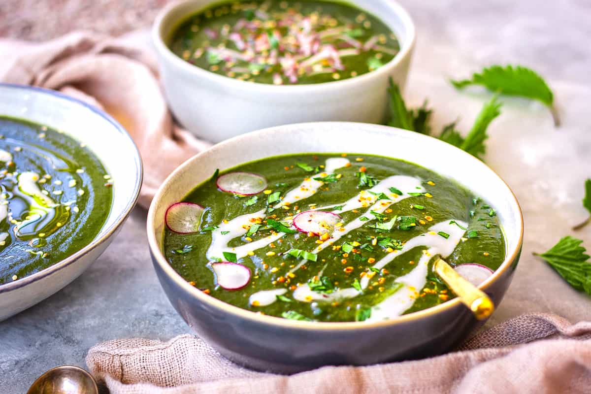 Close up image of three bowls of Nutrient-Rich Nettle Soup. The bowl in the foreground is decorated with yoghurt swirls, parsley, radish slices and seeds.