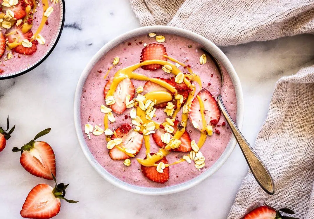 Overhead image shows a Strawberry Smoothie Bowl, decorated with strawberries, peanut butter and oats. A second bowl is visible, as is a linen napkin and some strawberry halves.