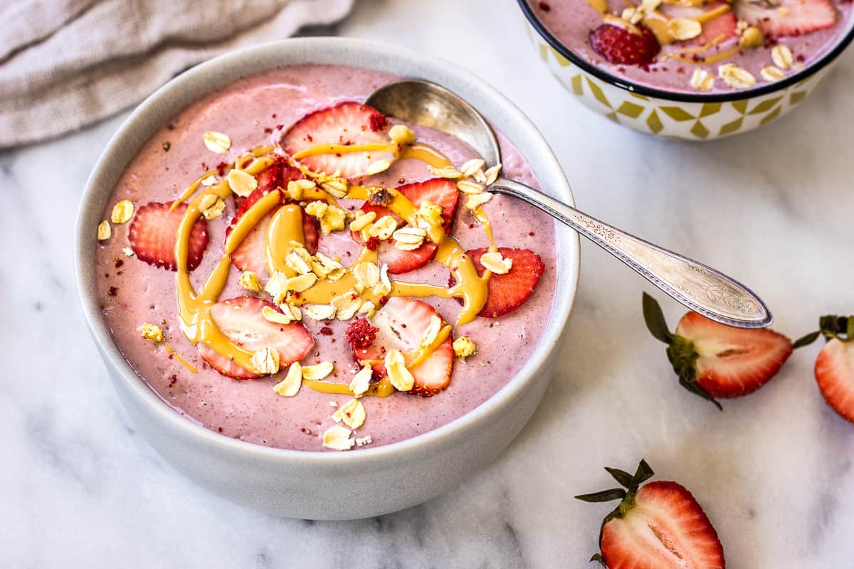 Image shows two Strawberry Smoothie Bowls, decorated with strawberries, peanut butter and oats. One bowl is in the foreground and we can see part of the other in the background. The bowls are surrounded by strawberries.