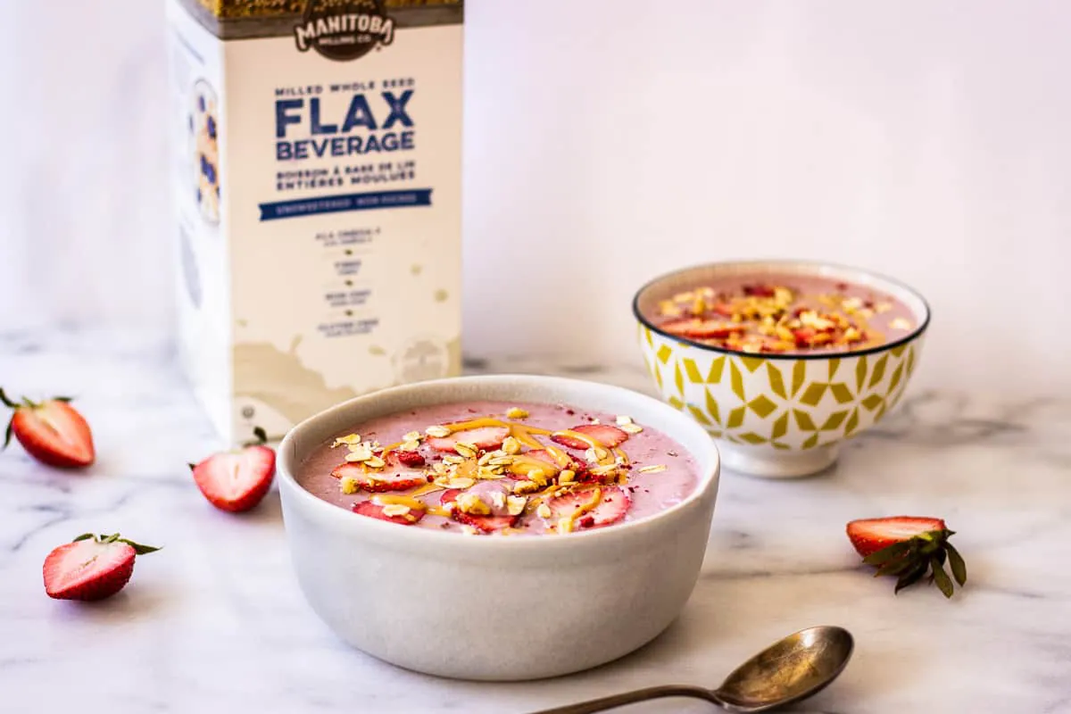Image shows two pink smoothie bowls on a marble surface in the foreground, decorated with strawberries, peanut butter and oats. In the background is a carton of Manitoba Milling Company Unsweetened Flax Beverage.