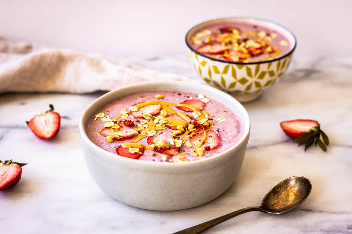Image shows two Strawberry Smoothie Bowls with Banana & Flax on a marble surface, decorated with strawberries, peanut butter and oats. Also visible are a tarnished silver spoon, some strawberries and a linen napkin.