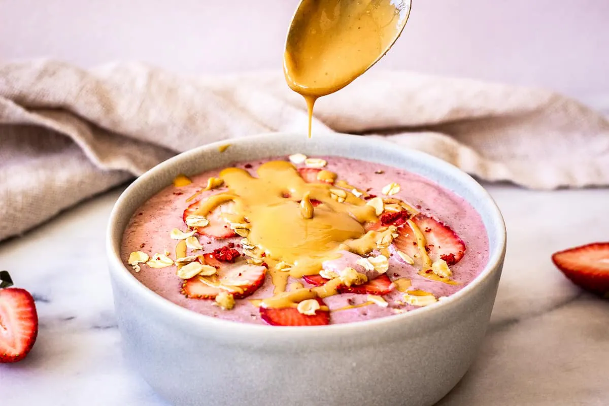Close-up image shows a pink smoothie bowl, decorated with strawberries, peanut butter and oats. A silver spoon is drizzling peanut butter onto the bowl.